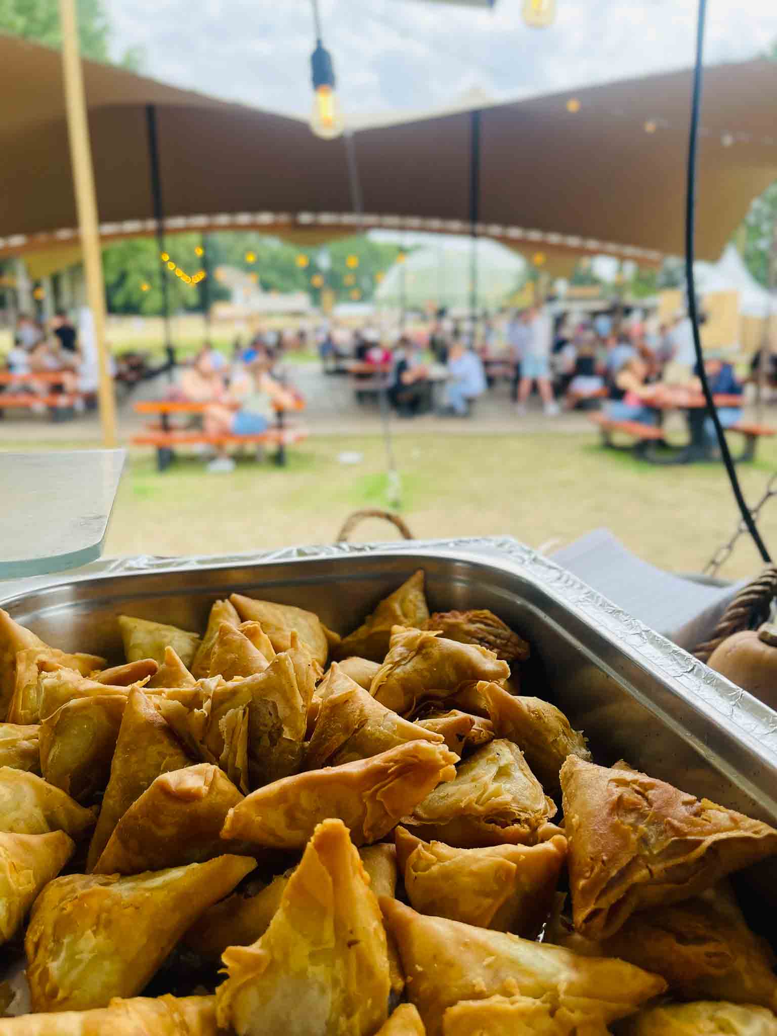In de voorgrond een schotel driehoekige gefrituurde pasta met een lekkere vulling met in de achtergrond bankjes waar mensen aan het eten zijn.